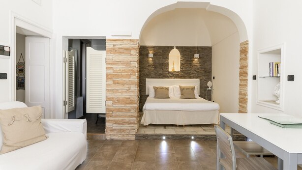 Un dormitorio con una pared de acento de piedra, una cama con ropa blanca, un arco blanco en la puerta y una pequeña mesa con una silla.
