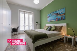 A bedroom with a bed, a desk, and a chair. There is a painting on the wall and a window with curtains.