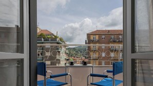 Un balcón con dos sillas azules y una mesa, con vista a una zona residencial con edificios y cielo despejado.