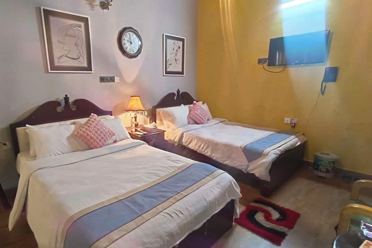 A hotel room with two beds, a nightstand with a lamp, a wall clock, and a television mounted on the wall.