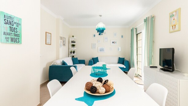 A modern living room with a white sofa set, a dining table with a decorative centerpiece, and a TV on a stand.