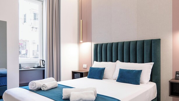 Una camera d'albergo con un letto, un comodino, una finestra con tende e vista sugli edifici.