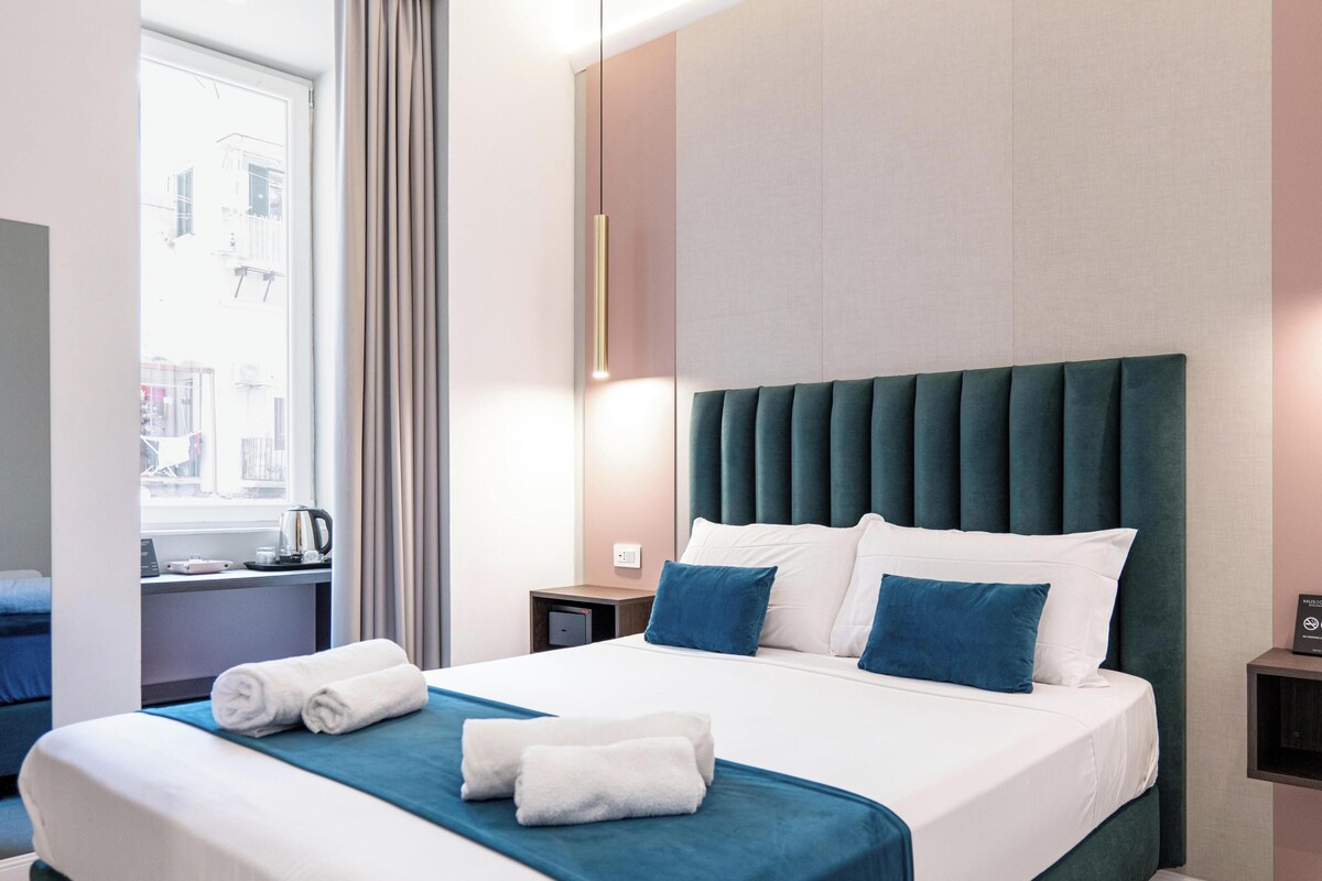 A hotel room with a large bed, a nightstand, a window with curtains, and a wall-mounted light fixture.