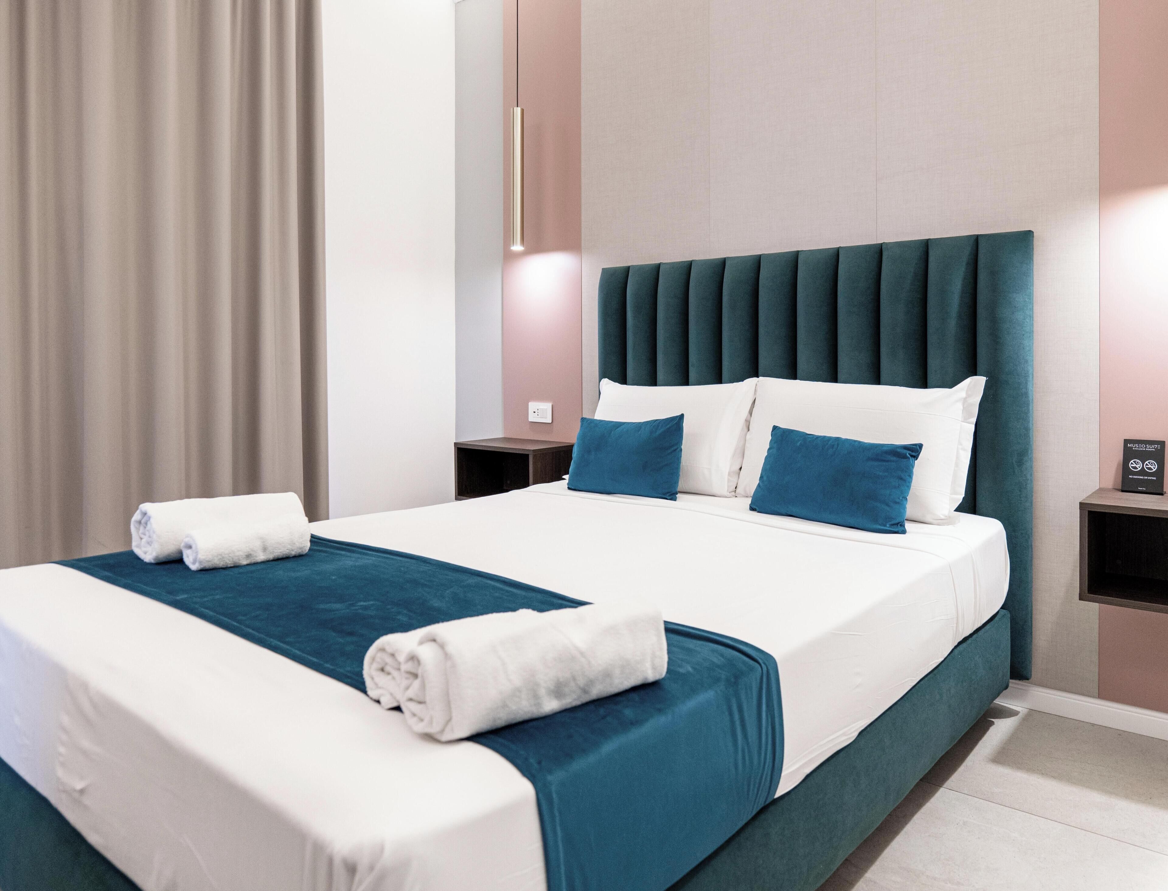 Quarto de hotel com cama, almofadas, mesa de cabeceira e luminária fixada na parede.