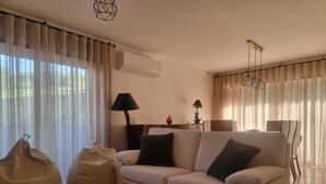 Uma sala de estar com um sofá bege, dois bean bags, uma mesa de centro e uma luminária. Há uma área de jantar com uma cadeira e uma mesa ao fundo.