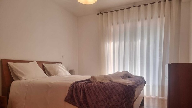 Quarto com cama, mesa de cabeceira, chão de madeira e janela com cortinas.