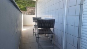 Pequena área de pátio murado com duas cadeiras de vime e uma mesa, paredes revestidas de azulejos, e vista para o verde lá fora.