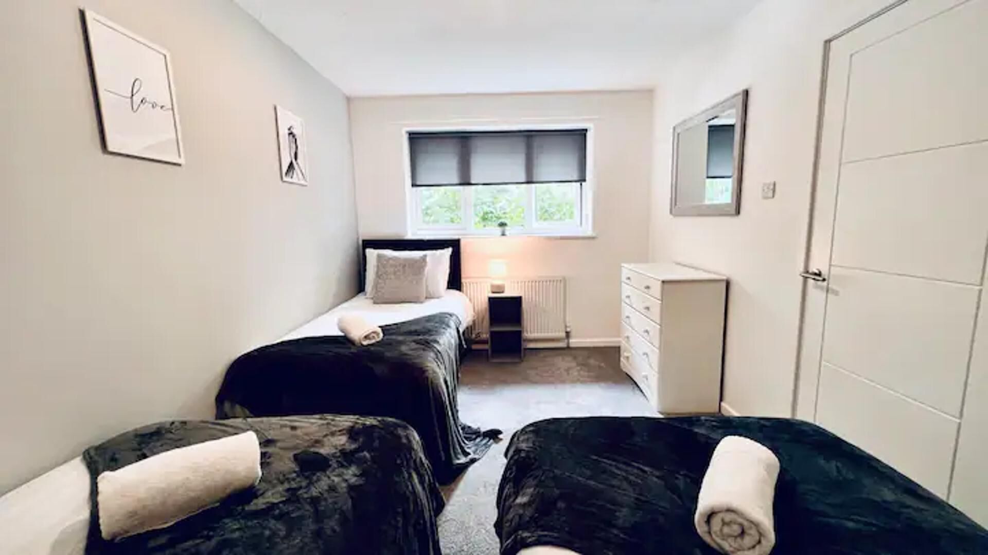 A bedroom with two beds, a dresser, a radiator, and a window with a view of greenery.