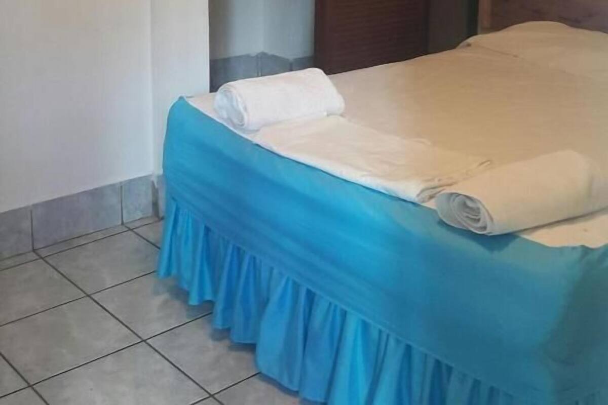 Un letto singolo con una gonna azzurra e biancheria da letto bianca, una porta in legno e un pavimento piastrellato.