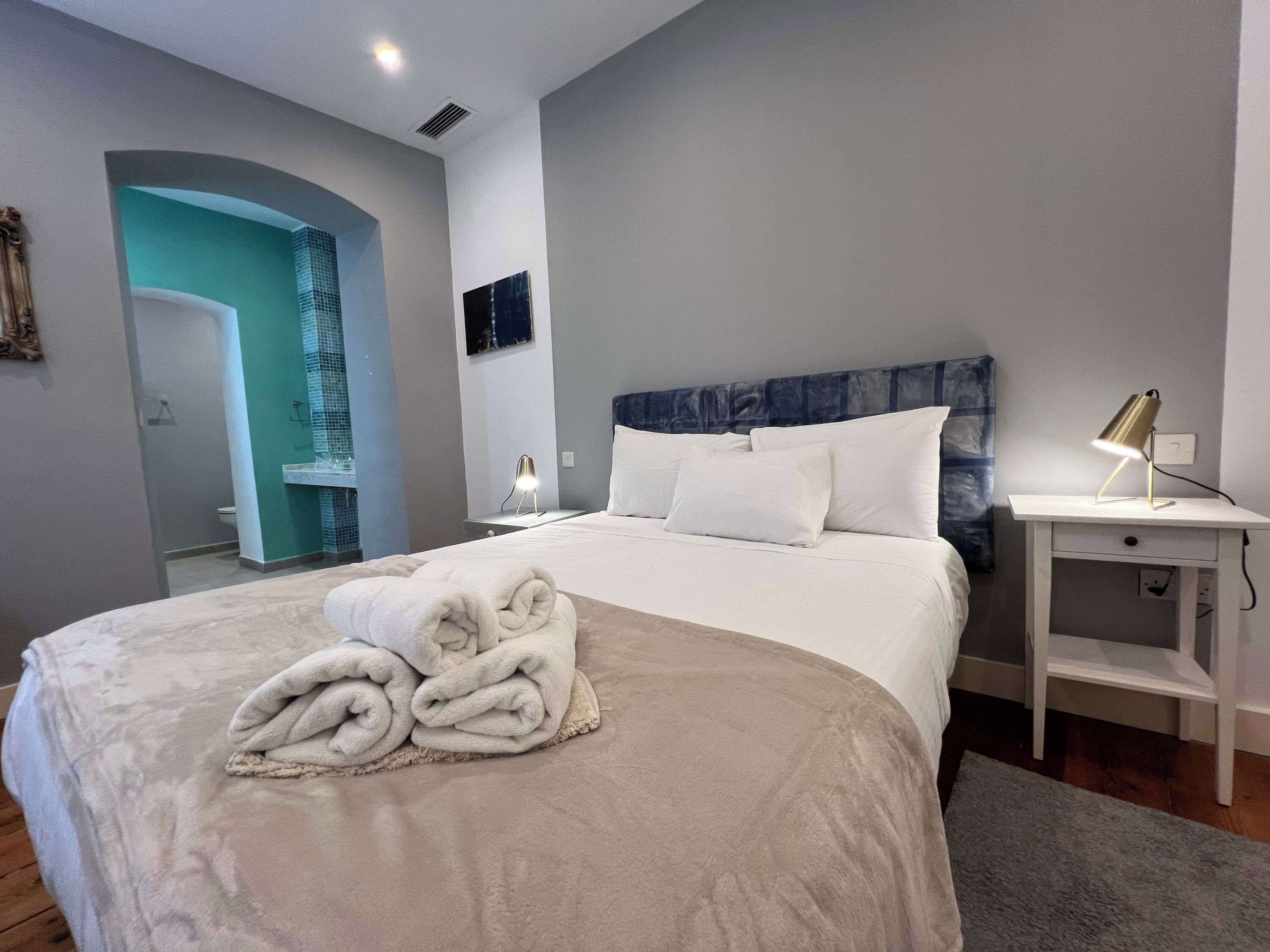Une chambre d’hôtel moderne avec un grand lit, une table de chevet avec une lampe, une salle de bain visible à travers une arche et une vue sur la ville.