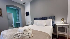 Une chambre d’hôtel moderne avec un grand lit, une table de chevet avec une lampe, une salle de bain visible à travers une arche et une vue sur la ville.