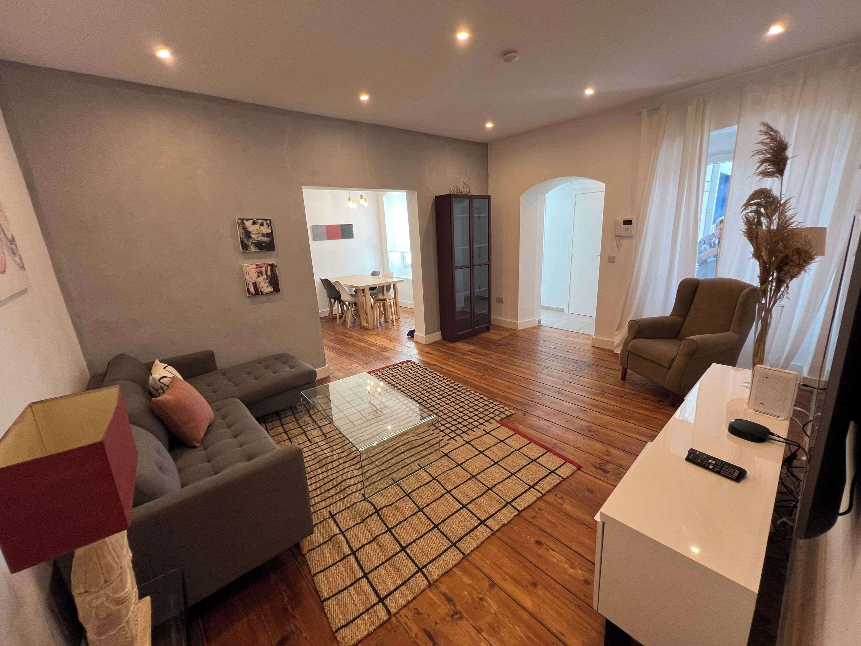 Un salon moderne avec un canapé gris, une table basse et un meuble pour la télévision. On aperçoit à l’arrière-plan un coin repas avec une table et des chaises.