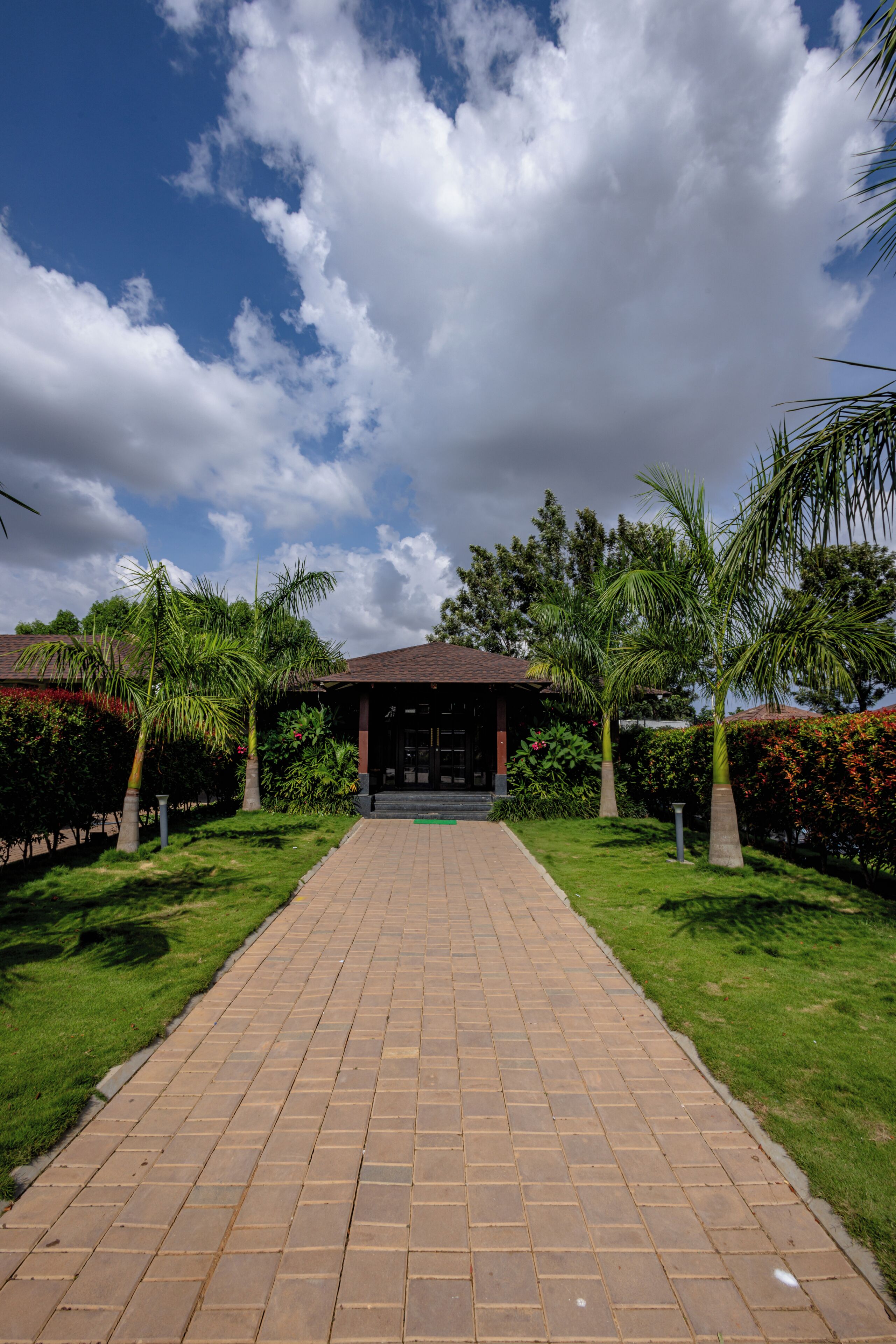 A paved pathway leading to a building with a dark roof, surrounded by greenery and palm trees.