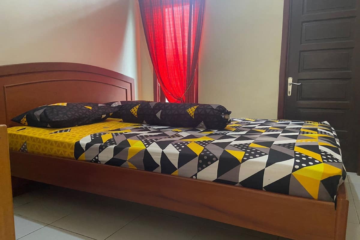 Una cama con una colcha estampada y almohadas, un cabecero de madera, una cortina roja y una puerta cerrada.