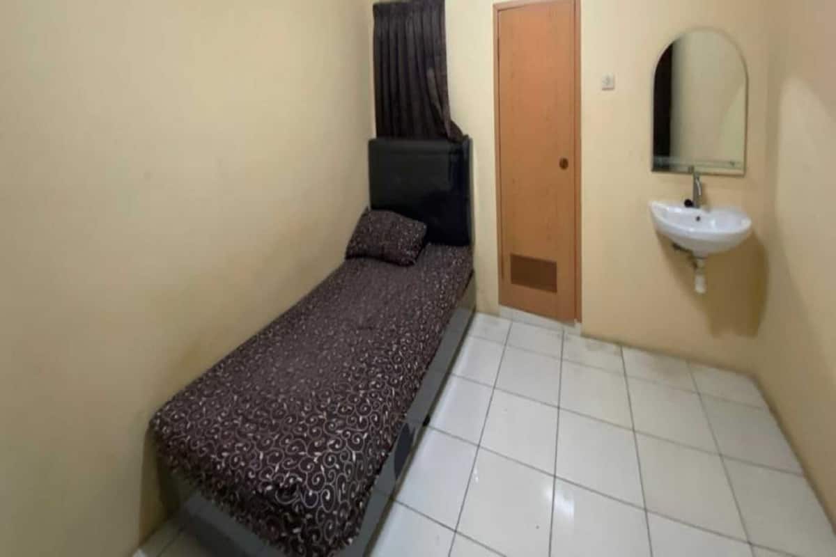 Una habitación pequeña con cama individual, colcha estampada, puerta de madera, lavamanos blanco y espejo en la pared.