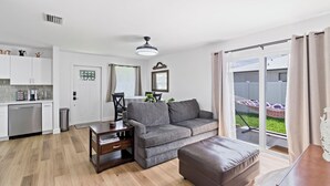 A modern living room with a grey sofa, a brown ottoman, and a dining area with a small table and chairs. A large window provides a view of the outdoors. - Cozy 2BD Near Ft. Myers Beach (Ft. Myers)