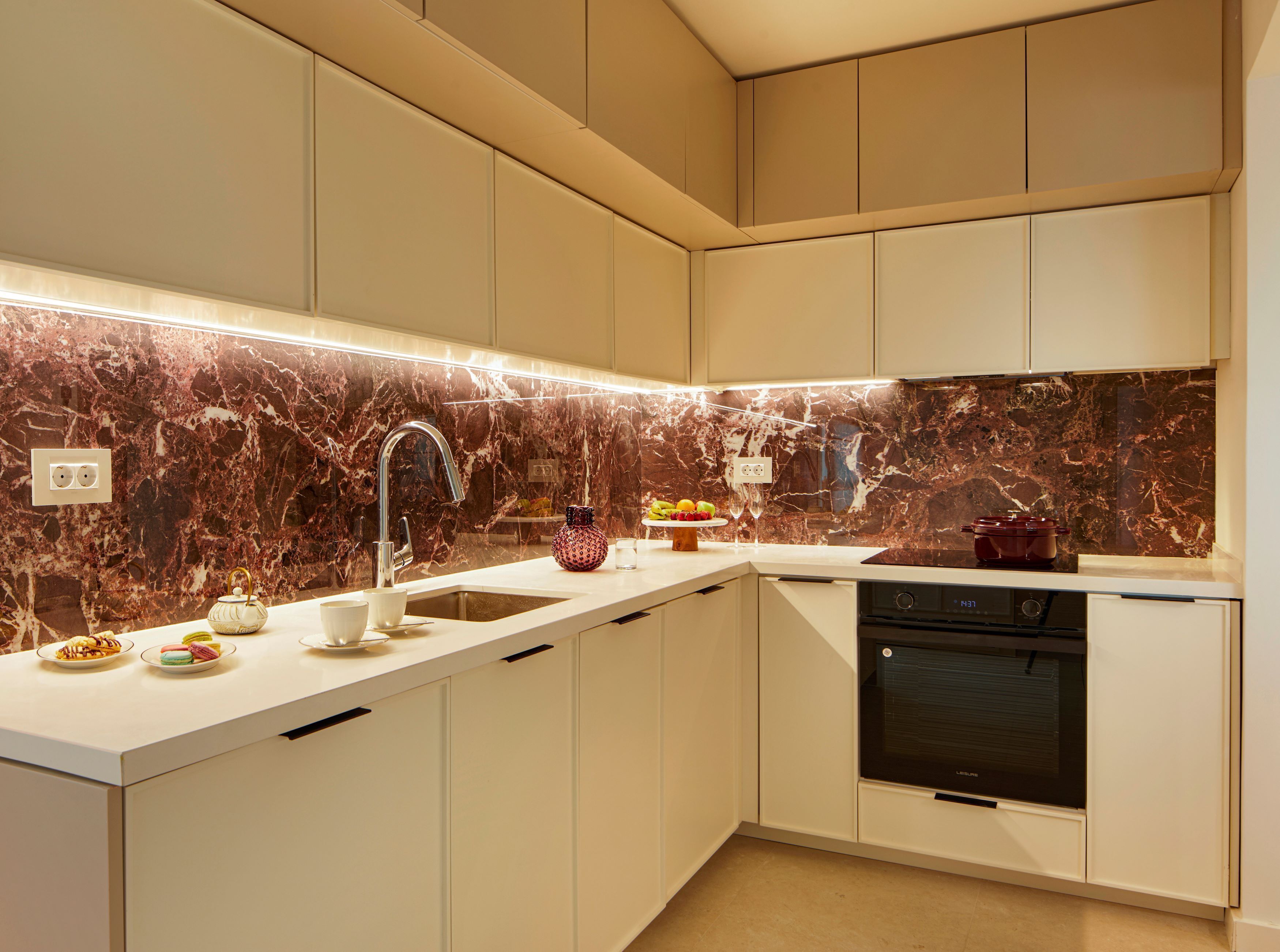 A modern kitchen with white cabinets, a marble backsplash, a stainless steel sink, and a built-in oven.