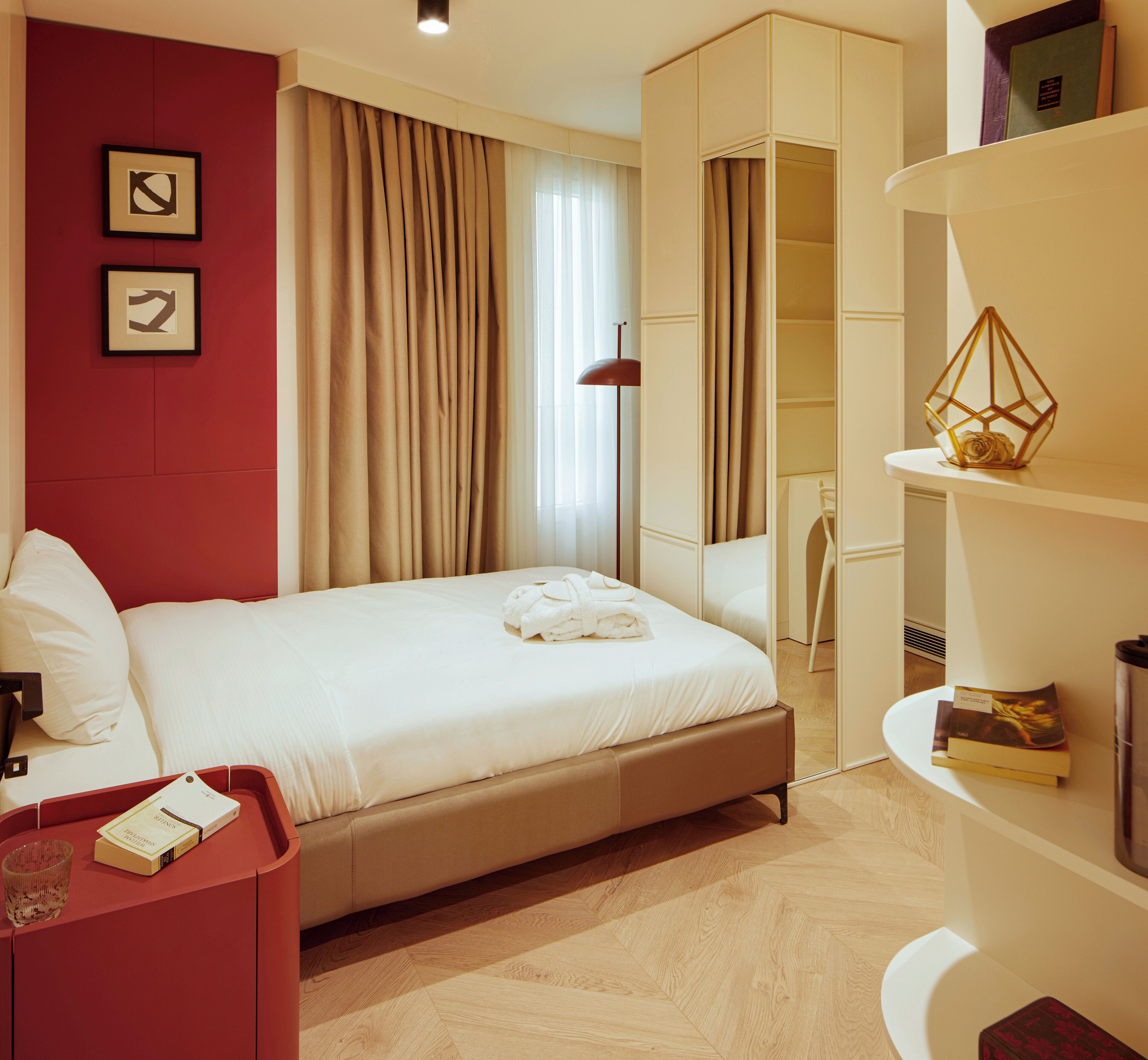A modern hotel room with a large bed, a red cabinet, a desk with a lamp, and a bookshelf.