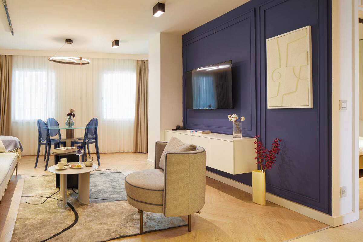 A modern living room with a sofa, a dining table with chairs, a flat-screen TV mounted on the wall, and a small side table with a vase.
