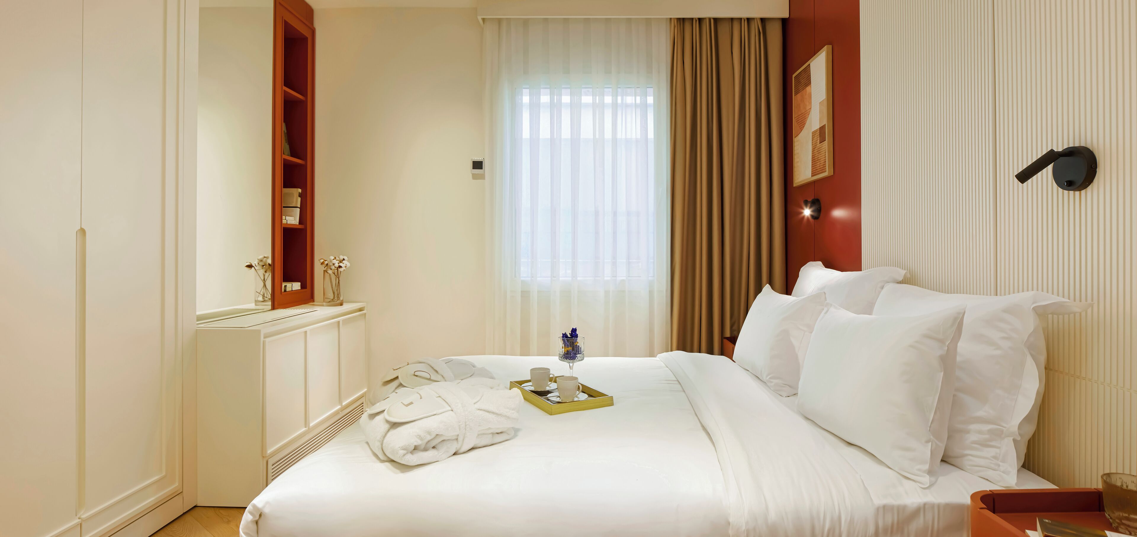 A hotel room with a neatly made bed, a bedside table with a tray of toiletries, a window with curtains, and a wall-mounted light fixture.