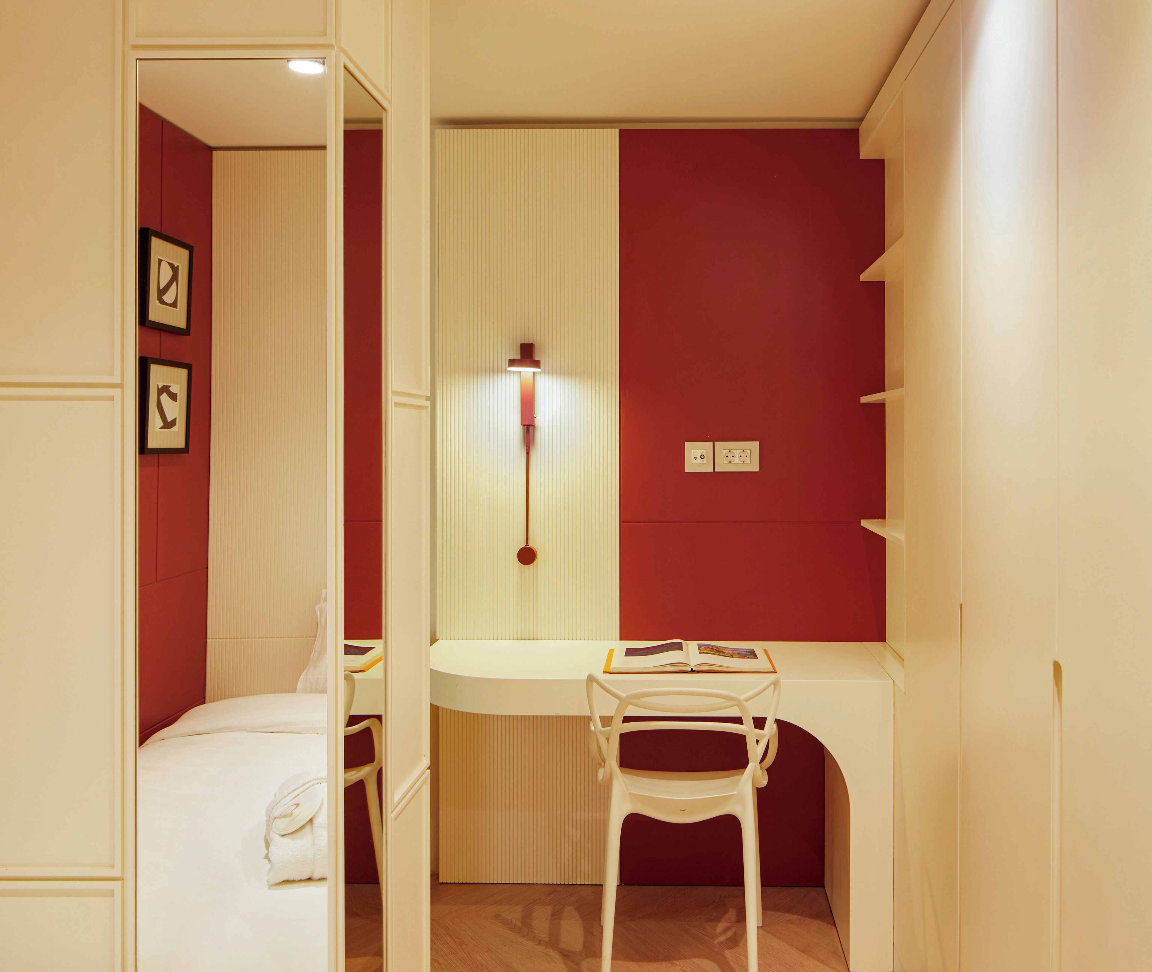 A compact room with a bed, a desk, and a chair. There is a wall-mounted light fixture above the desk.
