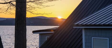 A house with a sloped roof and a window with a shutter, overlooking a body of water during sunset.