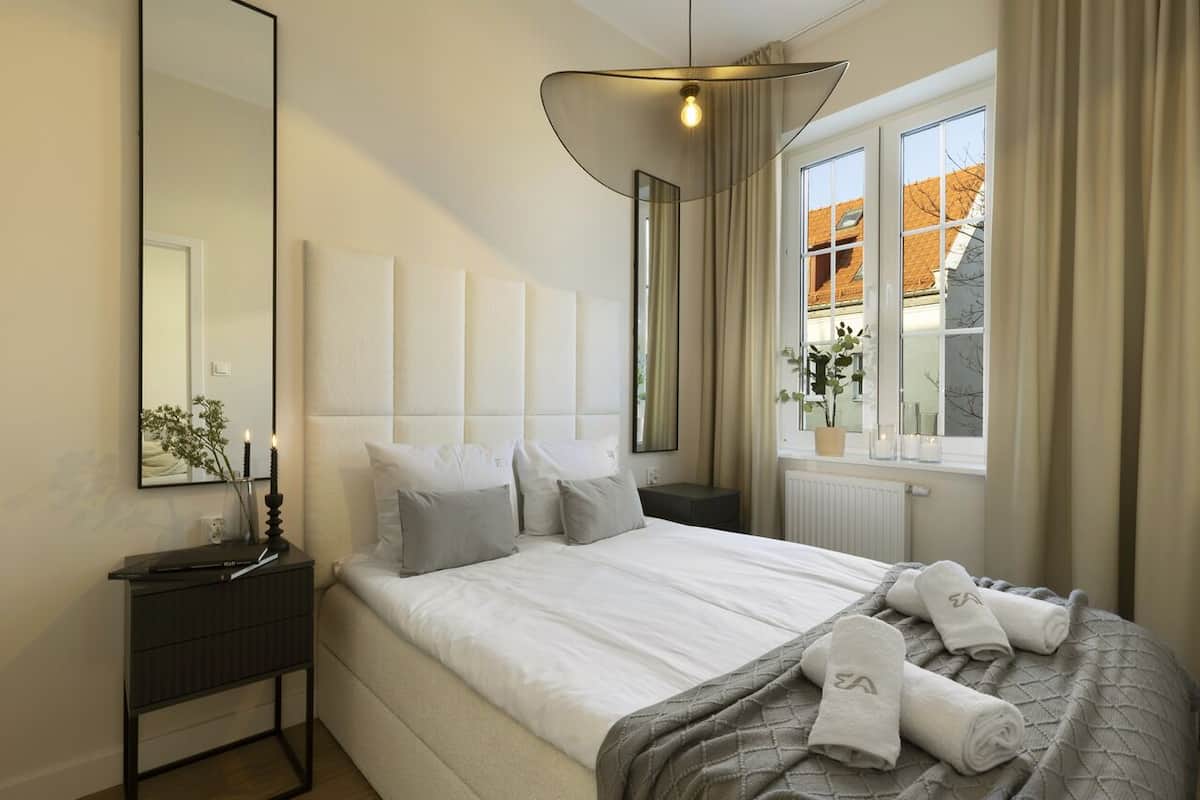 A modern hotel room with a large bed, a bedside table, a mirror, and a window with a view of buildings.