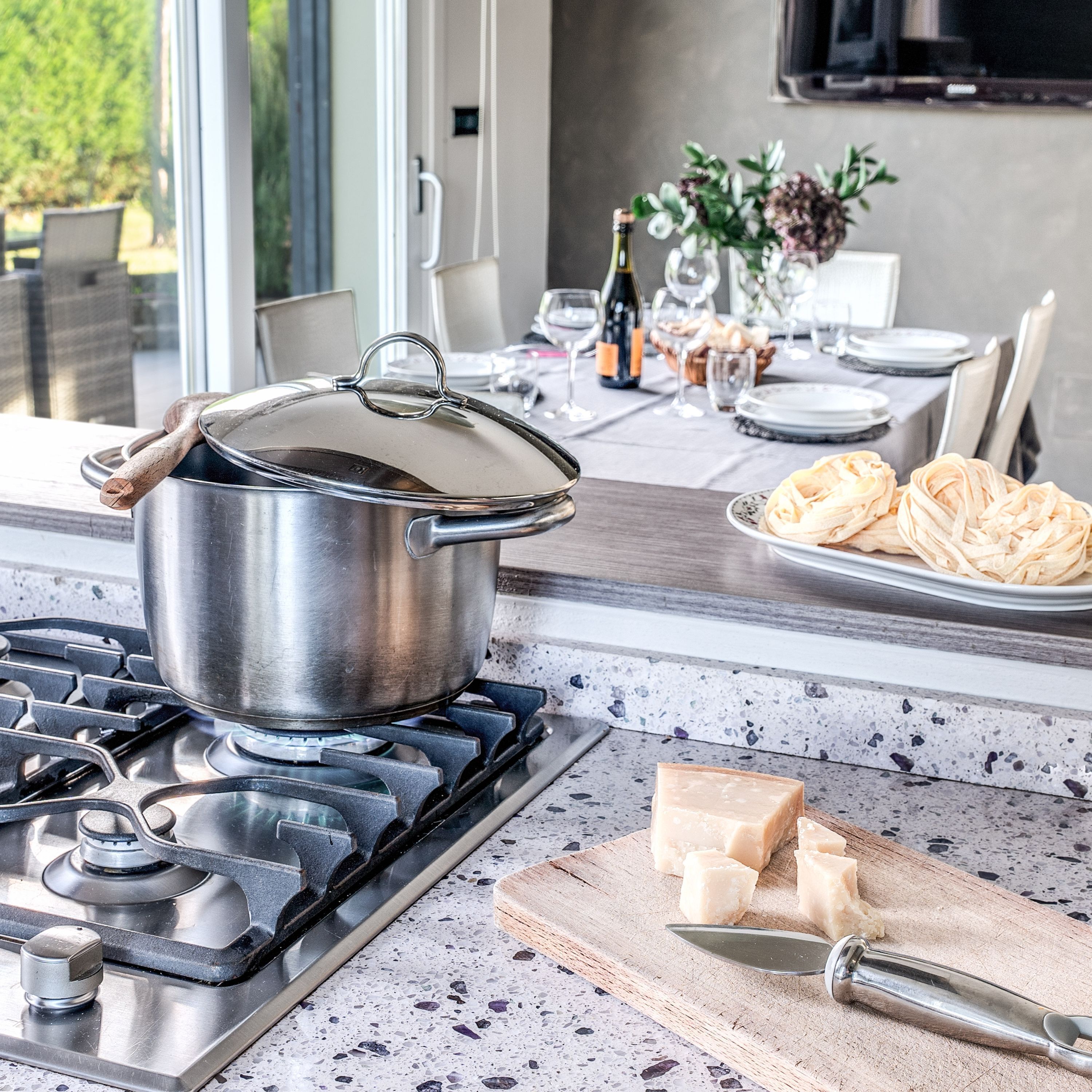 A kitchen with a stove, a pot on the burner, a cutting board with cheese, and a bottle of wine on the counter.