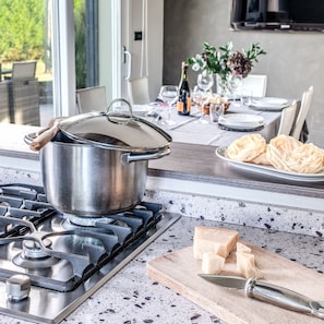 Une cuisine avec un fourneau, une casserole sur la plaque de cuisson, une planche à découper avec du fromage et une bouteille de vin sur le comptoir.