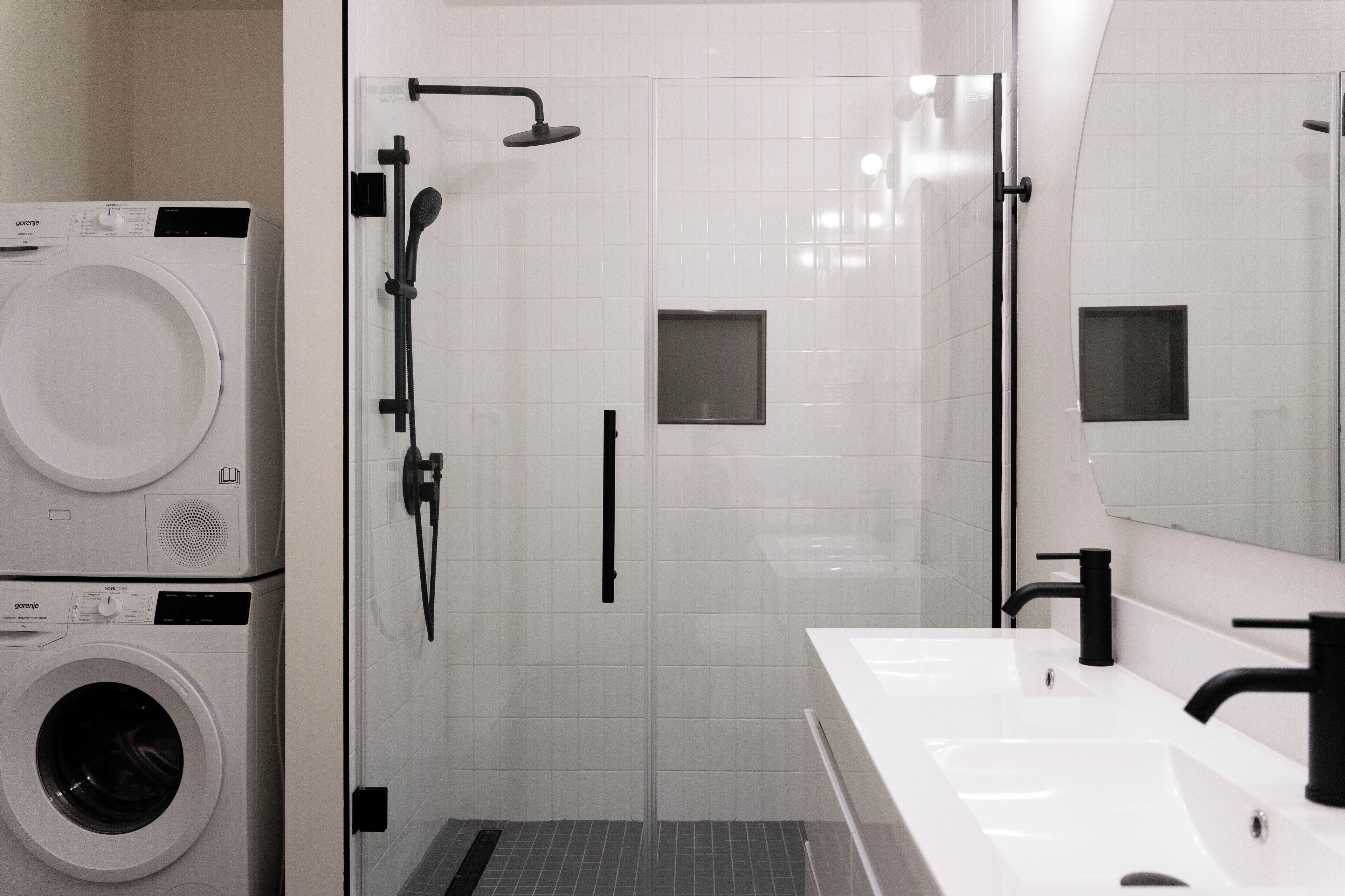 Une salle de bain moderne dotée d’une douche à parois de verre, d’un lavabo double avec des robinetteries noires, et d’un appareil combiné lave-linge et sèche-linge superposés.