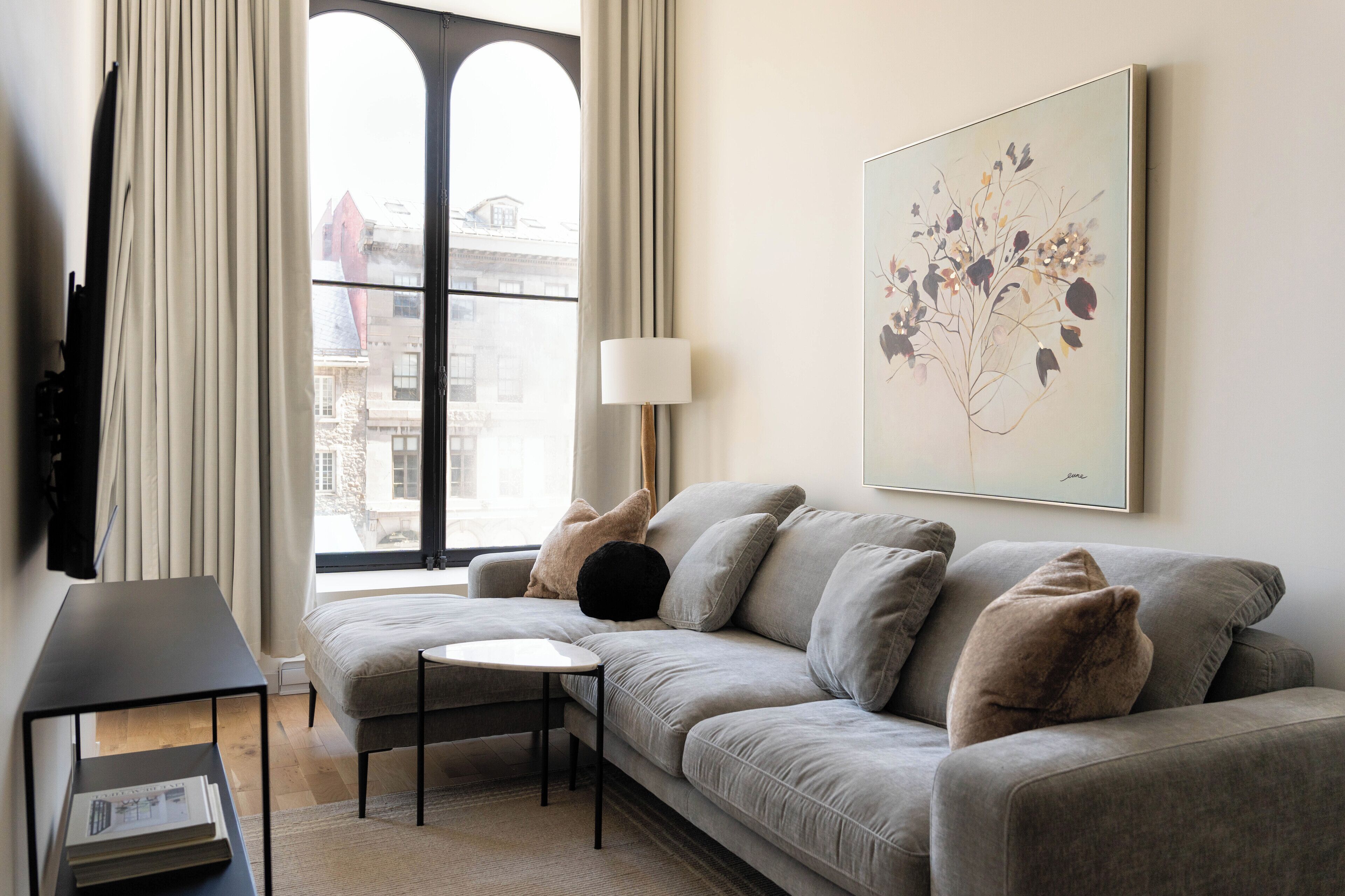A modern living room with a grey sofa, a small round table, and a lamp. There is a large window with curtains, a painting on the wall, and a view of buildings outside.