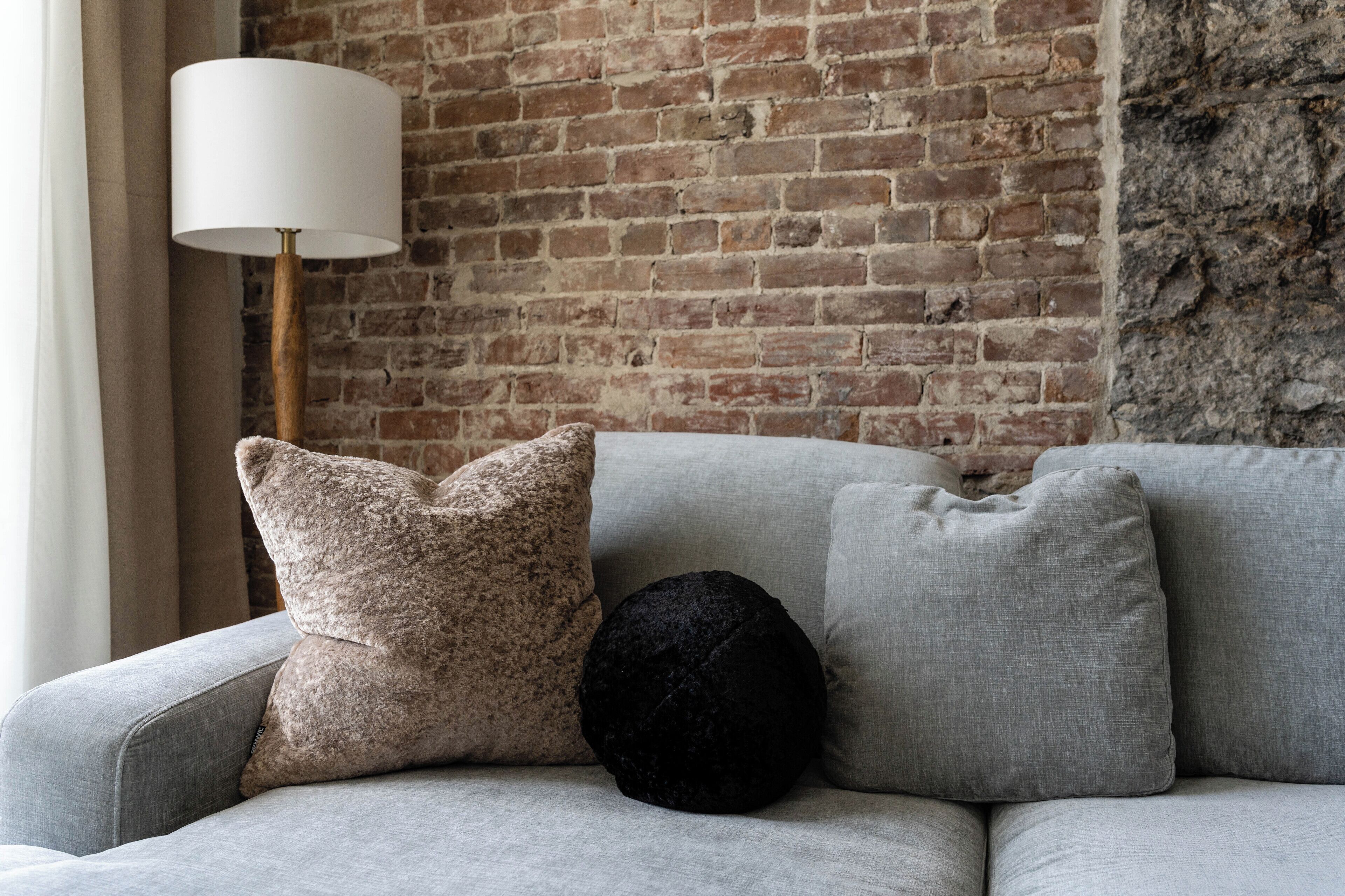 A grey sofa with three cushions against a brick wall.