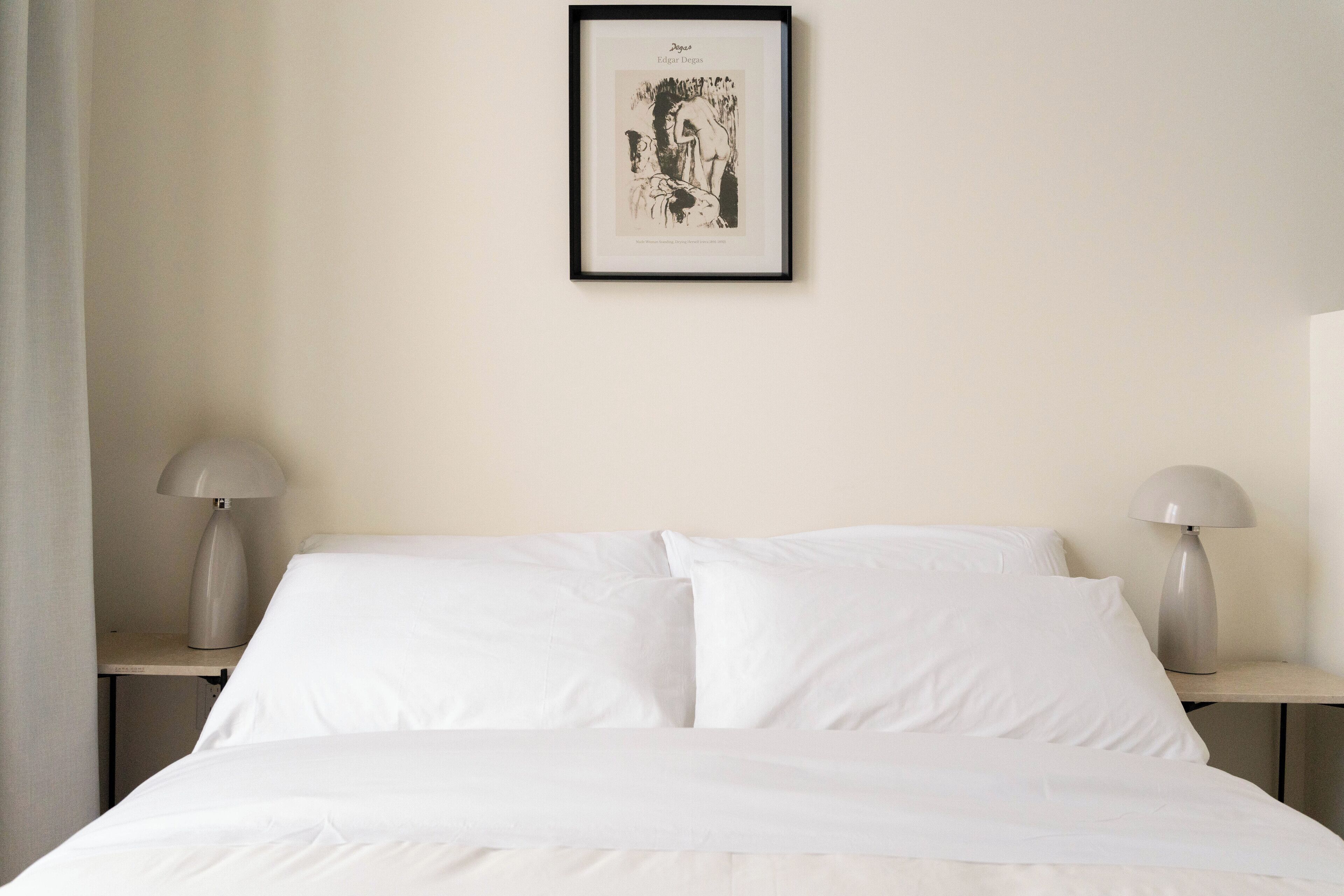 Un lit bien fait, avec du linge de lit blanc, deux lampes de chevet et une œuvre d’art encadrée au mur.