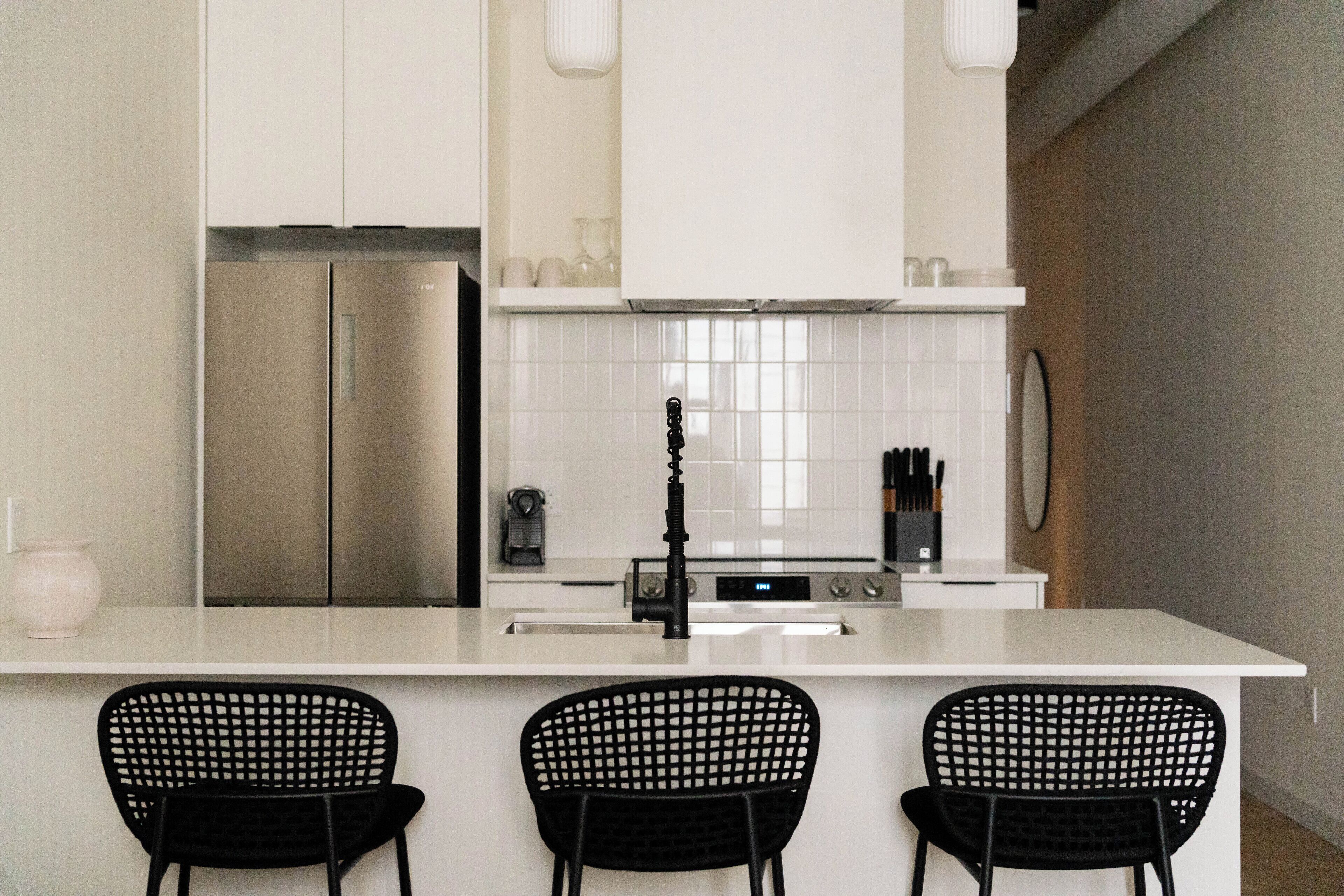 Cocina moderna con armarios blancos, un refrigerador de acero inoxidable, un grifo negro y tres sillas negras en la isla.