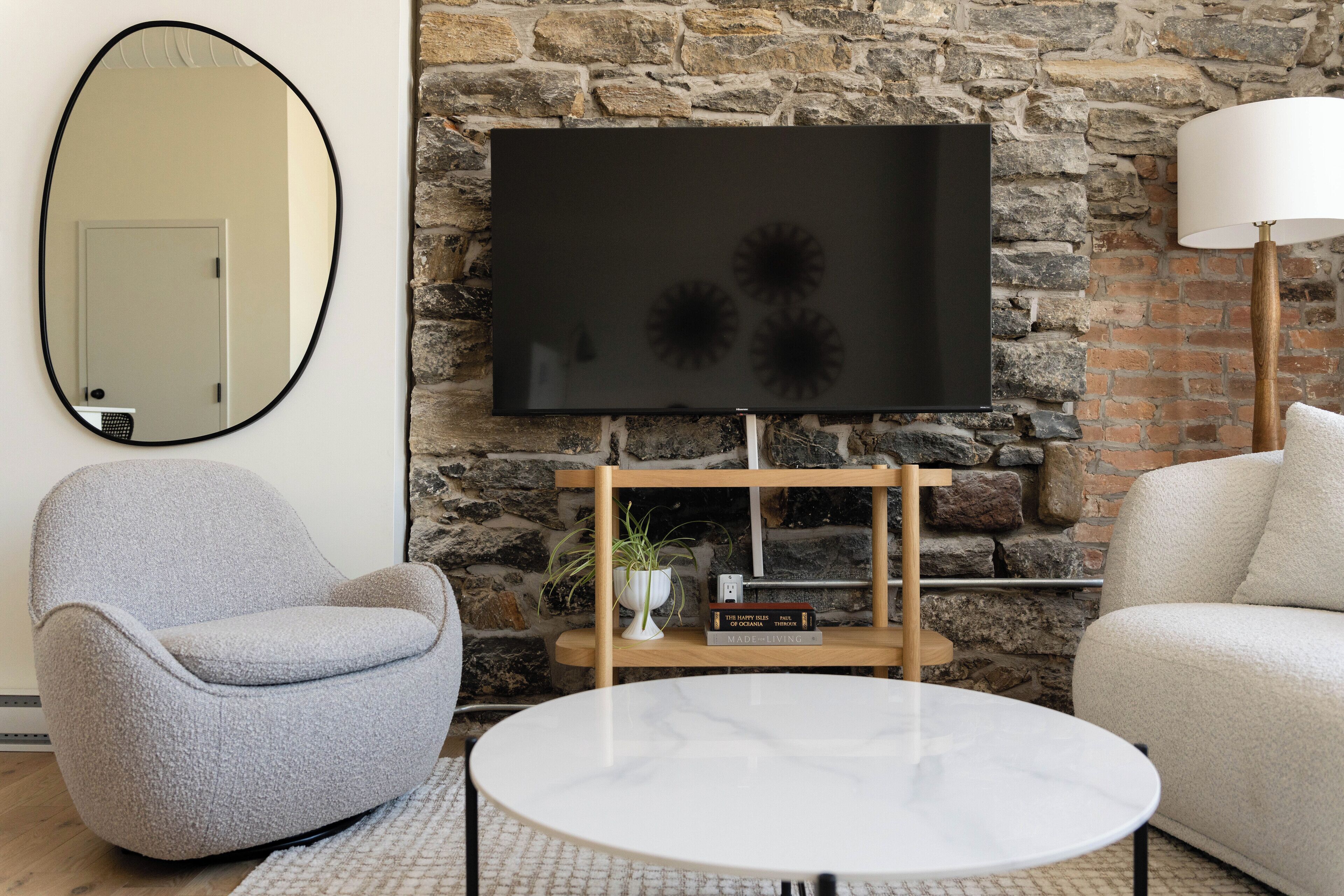 A living room with a stone wall, a flat-screen TV mounted on it, a round white table, and two armchairs.