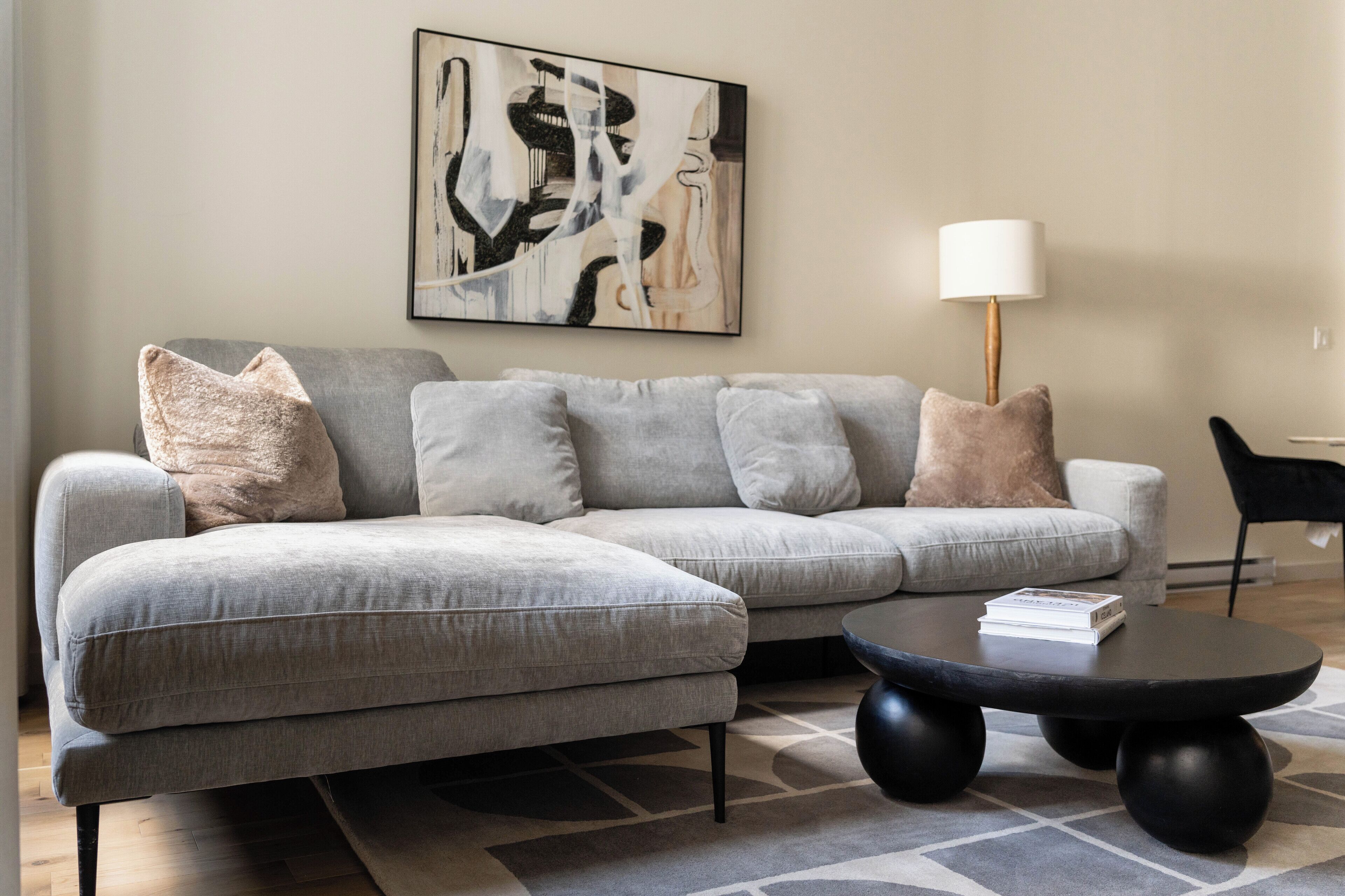 A modern living room with a grey sofa, multiple cushions, a black coffee table, and a painting on the wall.