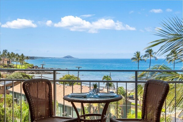 A balcony with wicker chairs and a small table, overlooking a beach and ocean.