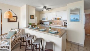 A modern kitchen with white cabinets, a central island with bar stools, a ceiling fan, and a view of the ocean through a mirror. - Summer Deal Oceanfront Kihei Penthouse with 32 Ft Lanai and Views (Kihei)