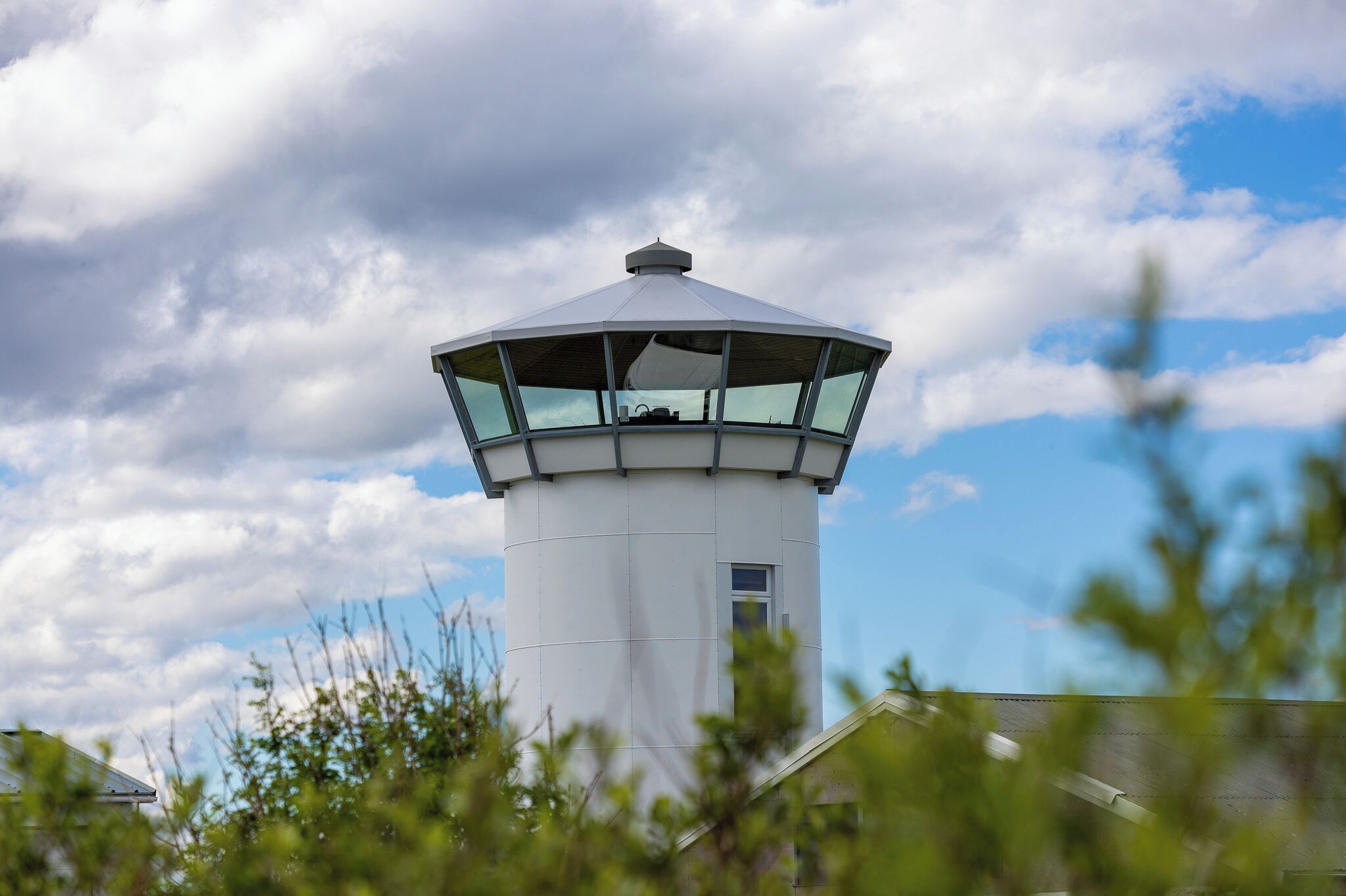 A lighthouse with a white cylindrical tower and a conical roof, surrounded by greenery.