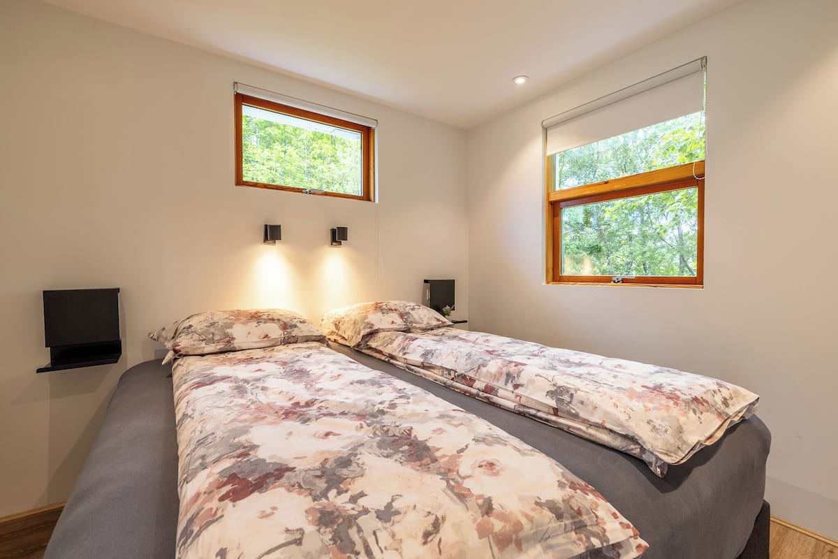A bedroom with two beds, two windows, a wall-mounted TV, and a shelf.