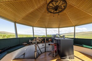 Espaço ao ar livre coberto com teto de madeira, balcão de bar, assentos e vista para colinas verdes.