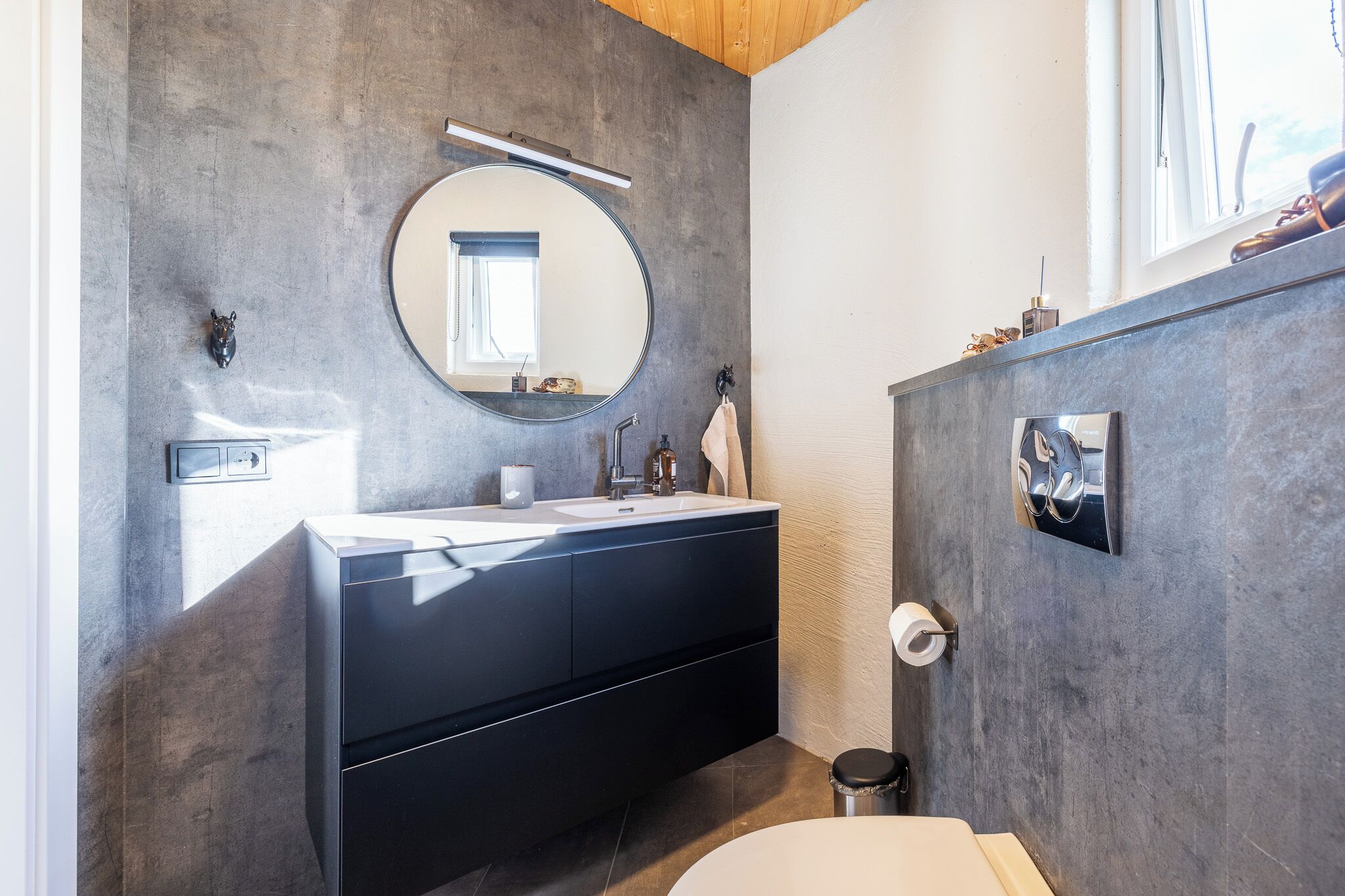 Un baño moderno con un espejo redondo, muebles oscuros y un inodoro a la vista.