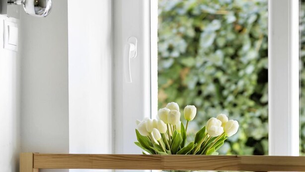 Een slaapkamer met een houten hoofdbord, een wit bed met een decoratief kussen en een vaas witte bloemen op de vensterbank.
