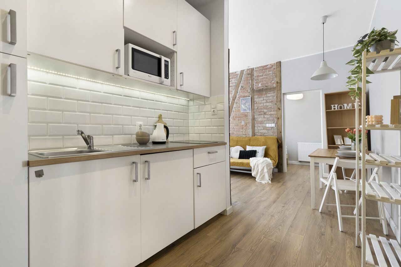 Una cucina moderna con mobili bianchi, un forno a microonde e un piano di lavoro in legno. C'è una zona pranzo con tavolo e sedie, e un letto con testiera bianca sullo sfondo.