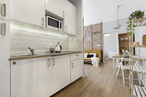 Eine moderne Küche mit weißen Schränken, einer Mikrowelle und einer Holzausziehtafel. Es gibt eine Essecke mit Tisch und Stühlen, und im Hintergrund ein Bett mit weißem Kopfteil.