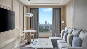 Habitación de hotel moderna con un televisor grande, una mesa de comedor redonda, un sofá con varios cojines y vistas a la ciudad a través de la ventana.
