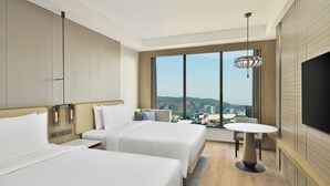 Habitación de hotel con dos camas, televisión, mesa de comedor y una ventana grande con vistas a la ciudad.