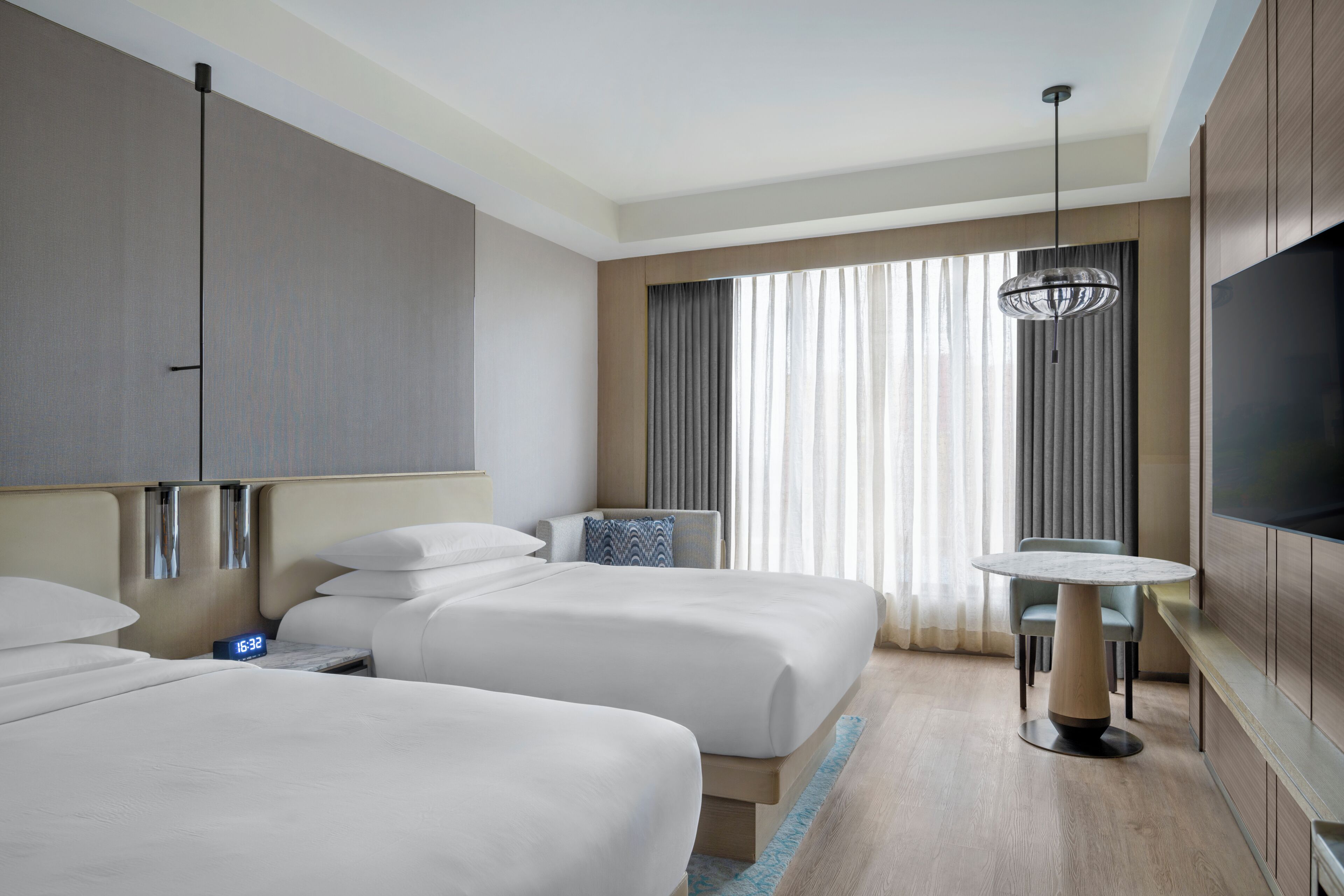 A hotel room with two beds, a TV, a dining table, and a large window with curtains.