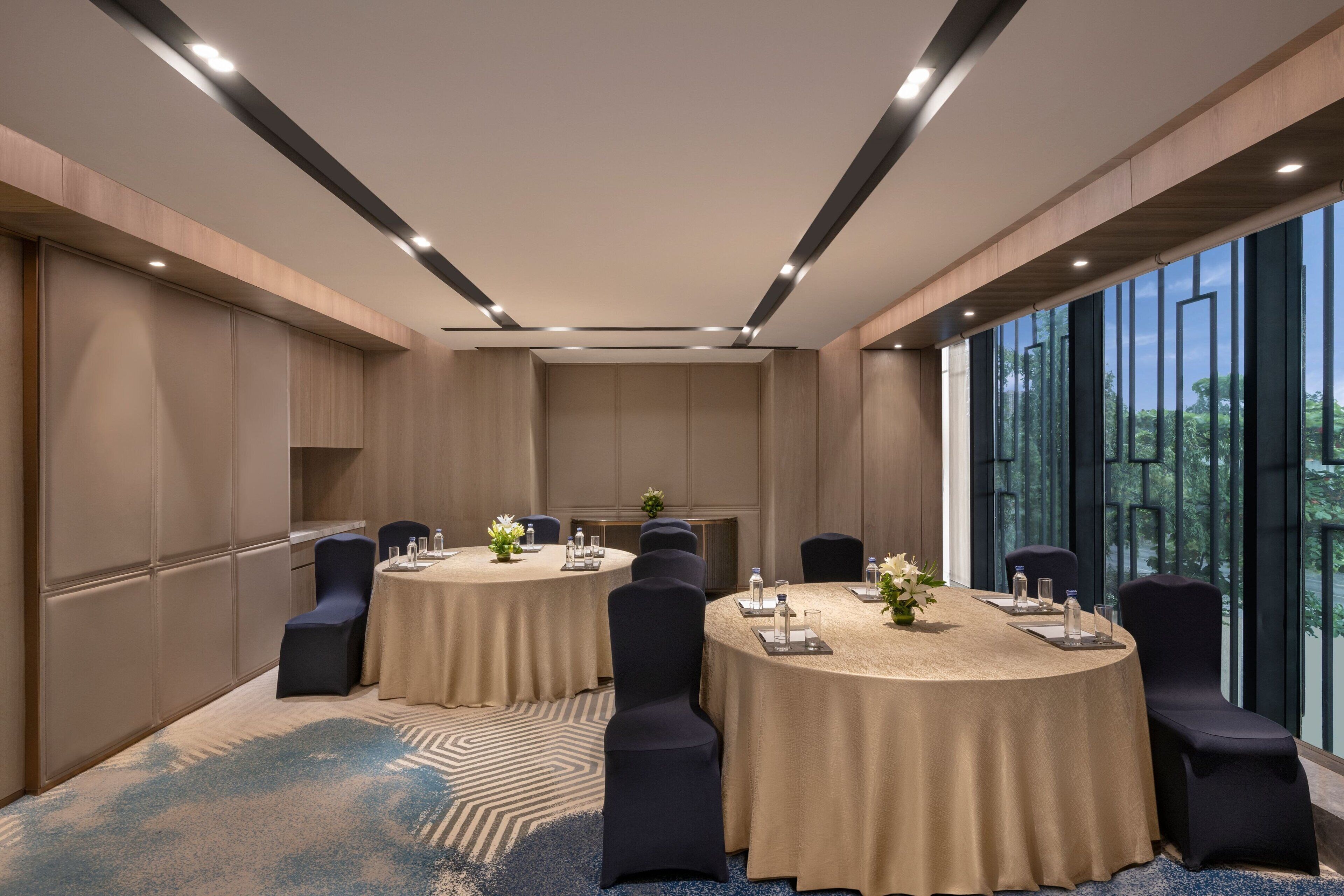 A conference room with two round tables, each surrounded by chairs, and large windows offering a view of greenery.