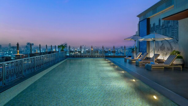 Una piscina infinita en la azotea con sillones de descanso y sombrillas, con vistas a un paisaje urbano al atardecer.